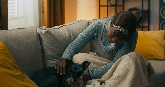 A young woman sits on the couch, gently embracing her dog after a morning walk. She looks at her pet with care and concern, enjoying the quiet companionship in their shared space.