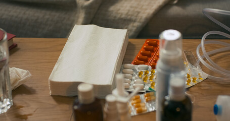 On the table lie paper handkerchiefs used for a runny nose, alongside various types of cold remedies including tablets, syrups, and nasal sprays arranged neatly in a sick room setting.
