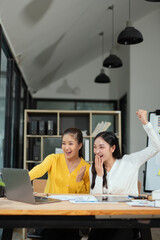 Two excited Asian businesswomen raising their arms and delighted at a successful company event in an office environment.
