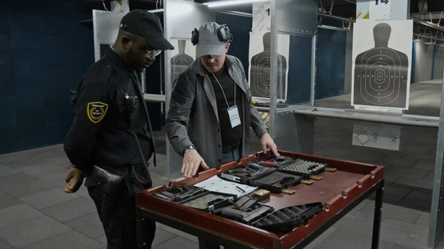 Medium full of young Caucasian male instructor demonstrating available guns at shooting range to African American trainee policeman in black uniform before marksmanship course