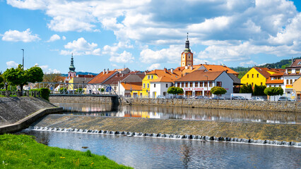Fototapeta premium The Otava River flows gently through Susice, showcasing quaint buildings with vibrant colors. Fluffy clouds float above, adding to the serene atmosphere on a beautiful day.