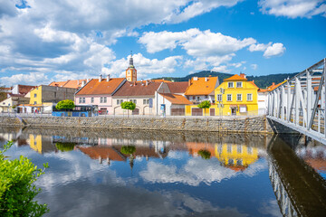 Obraz premium Sunlight reflects off the Otava River in Susice, Czechia, showcasing vibrant townhouses and a quaint bridge against a backdrop of blue skies and fluffy clouds.