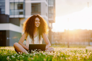 woman with laptop sits outdoors in sunlit grass, embracing digital nomad lifestyle. urban backdrop...