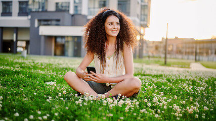 young woman with smartphone sitting cross-legged on green grass in urban park. smiling and enjoying sunset. casual outdoor lifestyle, urban relaxation, modern communication.