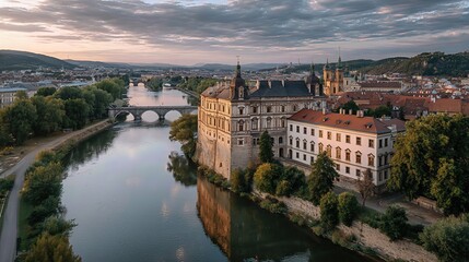 Obraz premium The majestic palace stands beside the river, framed by an urban backdrop and a bridge silhouetted against a cloudy sunset. 