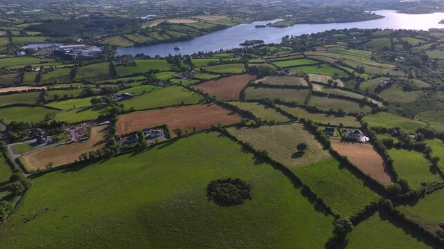 Lough Sillan, County Cavan, Ireland, June 2023. Drone establishing pullback above rolling farm fields sloping down to lake edge of water on sunny day, afternoon light casting long shadows
