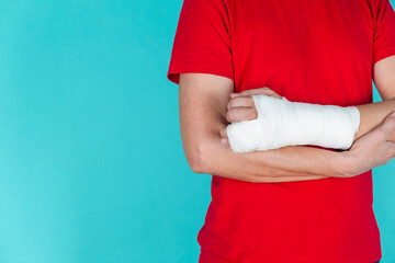Medical Concepts. Closeup of Body Parts of Caucasian Man in Red Shirt with Broken Hand In Plaster and Bandaid Isolated on Turquoise Background.