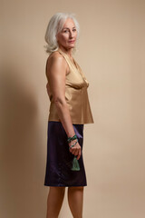 Ageing Concepts. Portrait of Senior Caucasian Blond Woman Posing in Casual Brown Blouse And Vilet Skirt In Studio Over Beige Background With Calm Facial Expression.