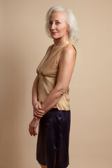 Portrait of Senior Caucasian Blond Woman Posing in Casual Brown Blouse And Vilet Skirt In Studio Over Beige Background With Crossed Hands Calm Facial Expression