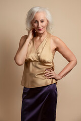 Natural Portrait of Senior Caucasian Blond Woman Posing in Casual Brown Blouse And Vilet Skirt In Studio Over Beige Background With Lifted Hand and Calm Facial Expression