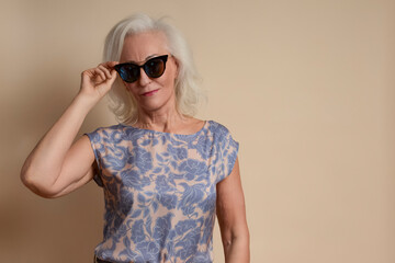 Tranquil Senior Caucasian Blond Woman Posing in Sunglasses in Casual Floral Blouse And Beige Pants In Studio Over Beige Background