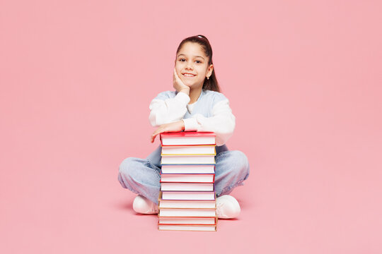 Full body young smiling happy school girl 8-9 years old wear blue casual clothes backpack sit hold stack of books isolated on plain pink background children portrait. Childhood kids education concept.