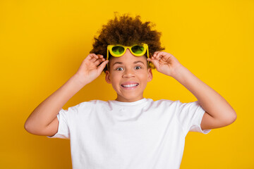 Cheerful boy wearing yellow sunglasses smiles in front of a vibrant yellow background, expressing...