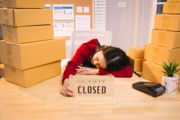 A middle-aged woman, who appears to be Asian, rests her head on a Sorry Closed sign amidst stacks...