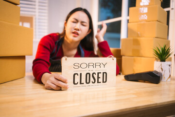 A middle-aged woman, who appears to be Asian, rests her head on a Sorry Closed sign amidst stacks...