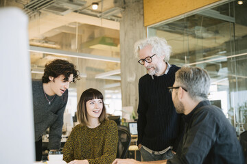 Midday in an open office space with neutral tones A multigenerational team is gathered around a desk, discussing ideas and sharing