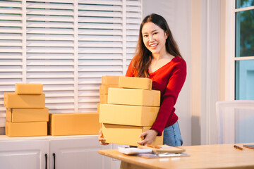 A smiling woman, who appears to be a middle-aged Asian individual, stacks cardboard boxes on a desk...