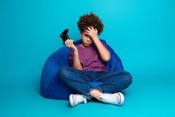 Young boy sits on a bean bag chair holding a game controller, appearing thoughtful against a vibrant blue background