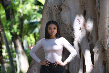 African woman, with long brown hair, young and beautiful, dressed in casual clothes with her hands on her hips next to the giant trunk of a ficus tree between sun and shadow. Beauty and fashion.
