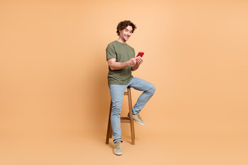Young man sitting on stool smiling while using a mobile phone on a warm beige background, wearing...