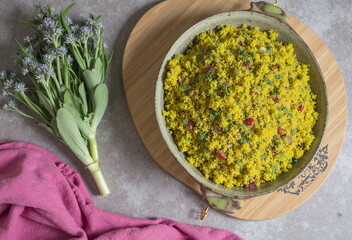 A vibrant yellow couscous salad with pomegranate seeds sits in a rustic bowl on a wooden tray, accompanied by fresh flowers. - Generative AI
