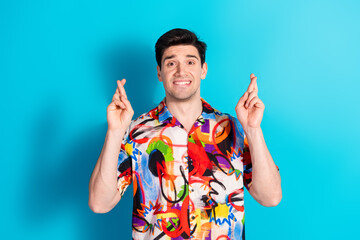 Young man in colorful shirt crossing fingers against blue background, expressing hope and optimism...