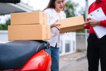 Attractive Asian delivery man in a red uniform hands a package to a middle-aged woman who is signing a clipboard next to a red motorcycle loaded with more boxes.
