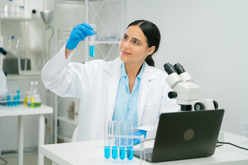 A scientist and researcher in laboratory examines test tube with blue liquid, surrounded by lab equipment like microscope, laptop, and test tubes. scientist researcher chemistry laboratory