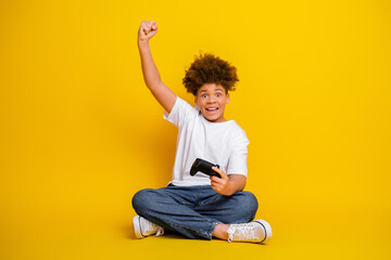 Fototapeta na wymiar Excited young boy with curly hair holding a gaming controller on a bright yellow background, celebrating enthusiastically