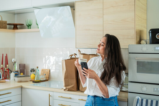 Young woman unpacking food packages in her kitchen and checking the list on her smartphone. Online food delivery.