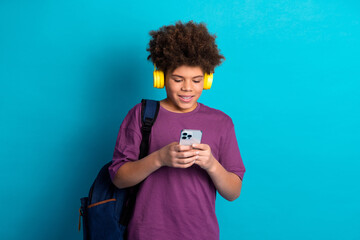 Teenage boy wearing headphones and using smartphone against blue background, enjoying leisure time, focused on device