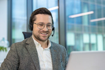 A close-up photo of a young man in a suit and headset sitting in an office at a desk and working on...