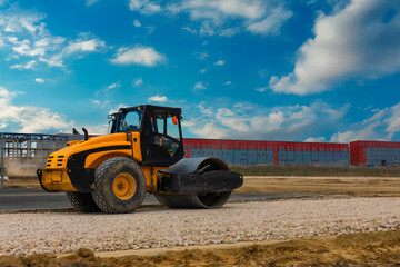 Yellow road roller compactor working on gravel at a construction site with a red and gray industrial building in the background.