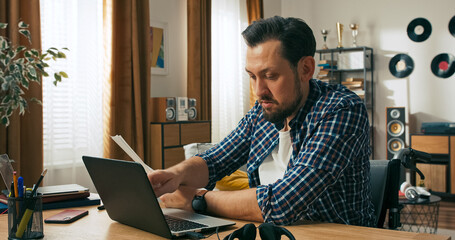 A young bearded brunette man sits at a table in front of a laptop, holding papers in his hands. He works remotely, checking information and creating graphics for a digital project.