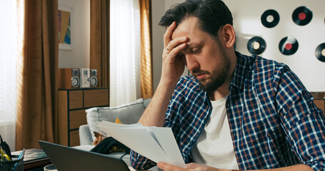 A sad and focused brunette man stares at overdue bills with concern. He is tired and mentally...