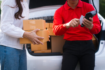 A delivery man, appearing to be Asian, in a red uniform hands packages to a middle-aged woman next to an open car trunk. She is paying with a credit card, paying for the delivered packages.