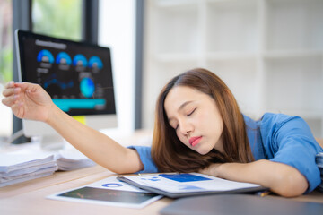 Young Asian office worker sleeping on desk after working on financial data analysis, showing exhaustion and stress from overwork in a modern office environment