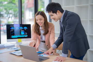 Two young professionals engaged in a collaborative business discussion, analyzing data on a laptop in a modern office setting. A computer screen displays charts and graphs in the background