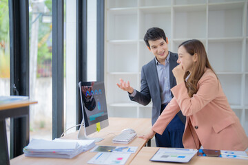 Two asian businesspeople discussing over desktop computer showing charts and graphs on the screen, analyzing financial data and planning project strategy in modern office