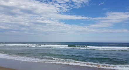 Fototapeta premium Ocean waves rolling onto a sandy beach under a cloudy sky for travel and relaxation themes.
