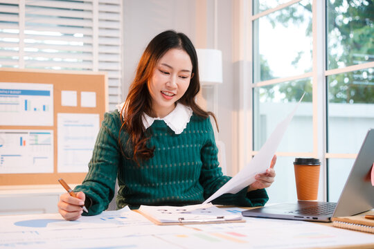 Confident middle-aged Asian professional works on a laptop amidst scattered charts and documents in a bright home office, a coffee cup nearby hinting at a dedicated work session.