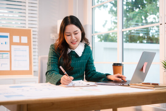 Confident middle-aged Asian professional works on a laptop amidst scattered charts and documents in a bright home office, a coffee cup nearby hinting at a dedicated work session.