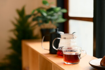 A glass filled with black coffee beside a pour over carafe on a wooden table with warm morning light