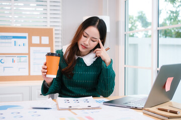 Confident middle-aged Asian professional works on a laptop amidst scattered charts and documents in a bright home office, a coffee cup nearby hinting at a dedicated work session.