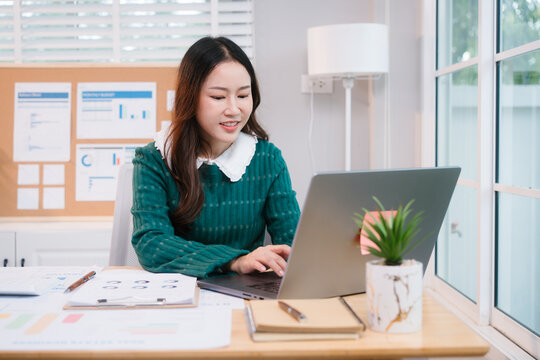 Confident middle-aged Asian professional works on a laptop amidst scattered charts and documents in a bright home office, a coffee cup nearby hinting at a dedicated work session.