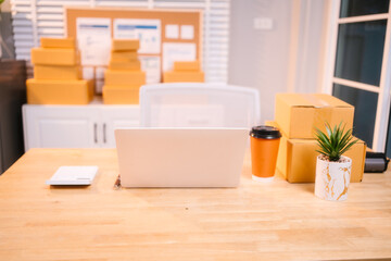 A neat workspace with a white laptop, stacked delivery boxes, a coffee cup, and a potted plant on a wooden desk, indicating a busy day of online sales packaging and fulfillment.