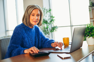Charming mature woman in a cozy blue sweater working on her laptop at home during daylight in a...