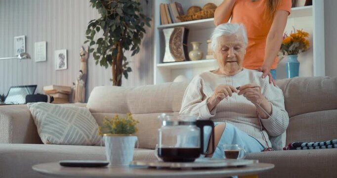 Mature daughter visits her old 80s mother, who knitting woolen socks sitting on the sofa at home in the living room.