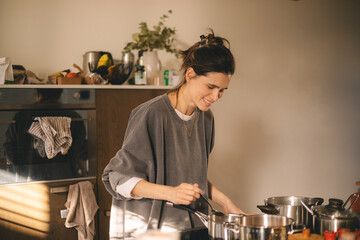 Beautiful happy young woman is cooking in the home kitchen and testing some soup from the pan on the stove. Cooking woman in kitchen stirring in pot making food for dinner. Young housewife smiling.