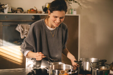 Beautiful happy young woman is cooking in the home kitchen and testing some soup from the pan on the stove. Cooking woman in kitchen stirring in pot making food for dinner. Young housewife smiling.